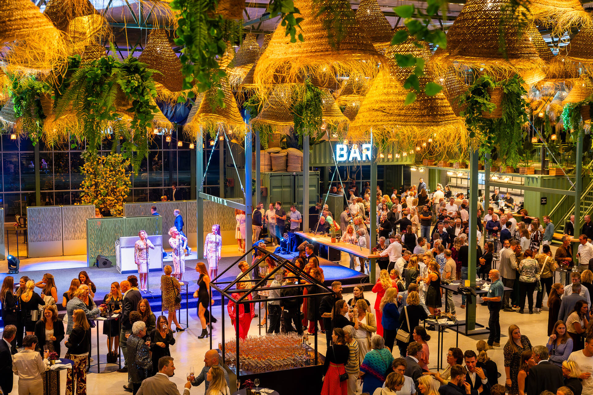 Foto van een evenement in de Evenementenkas van de Midden Nederland Hallen waar drie zangeressen optreden op een podium en bezoekers erom heen staan te borrelen en kletsen.